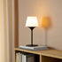 Zwarte Ambience lampvoet met witte lampenkap op een houten boekenkast met boeken voor warme sfeerverlichting in een modern interieur van sackit