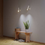 Twee witte Expo wandlampen geven gericht licht op een grijze muur boven een houten bank met plant en schoenen in een warm modern interieur