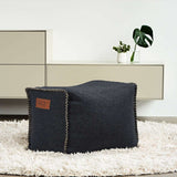 Donkergrijze vierkante poef van sackit in een modern interieur met een zacht wit vloerkleed en een plant op de achtergrond