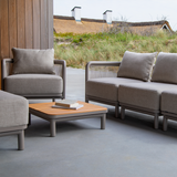 Modulaire grijze loungesofa van sackit met bijpassende fauteuil en houten salontafel op een overdekt terras met duinen op de achtergrond