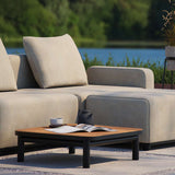 Outdoor salontafel van zwart gepoedercoat aluminium met teakhouten blad voor een beige loungesofa bij het water met tijdschrift en kopje
