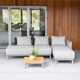 Lichtgrijze modulaire loungesofa met chaise longue en bijpassende pouf van sackit op een overdekt terras met houten wand en lage tafel met plant