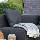 Donkergrijs Cobana kussen op een weerbestendige loungesofa voor buiten met een zachte gestreepte plaid en decoratieve lantaarns in de tuin