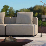Beige buitenkussen op een modulaire loungesofa van sackit op een zonnig terras met groen op de achtergrond en een stijlvolle plantenstandaard naast de bank