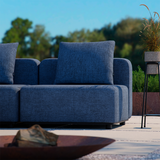 Donkerblauwe buitenbank met zachte rug en zitkussens op een modern terras met planten en een zonnige groene omgeving ontworpen door sackit