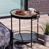 Zwarte metalen serveertafel voor buiten met rond houten blad en afneembaar dienblad met koffie en ontbijt naast een loungesofa op een zonnig terras