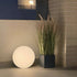 sackit Light 350 ronde buitenlamp met warm wit licht naast een plantenbak op een terras voor sfeervolle verlichting tijdens lange avonden