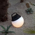 sackit Light 250 draagbare buitenlamp met handvat geeft warm wit licht in een grindtuin tussen siergrassen en vetplanten. De ronde lamp is waterbestendig.