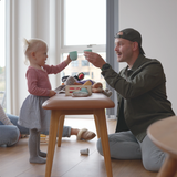 Kind en vader spelen samen aan een rond houten meubel in een lichte woonkamer met speelgoed en knuffels terwijl ze elkaar lachend aankijken