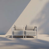 Lichtgrijze Kirra loungesofa voor twee personen van sackit met diepe kussens en armleuningen in een minimalistische buitenruimte met zachte schaduwen