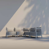 Modulaire grijze loungesofa voor buiten met hoekopstelling zachte kussens en gevlochten zijpanelen tegen een lichte wand met schaduwen van bladeren van sackit