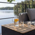 Teakhouten opbergtafel voor buiten naast een grijze loungesofa met glazen kan en drinkglazen op een terras aan het water