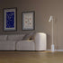 sackit vloerlamp met slank wit aluminium profiel en marmeren voet naast een moderne grijze bank in een stijlvol interieur met wandkunst
