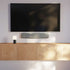 Slanke grijze soundbar onder een zwart televisiescherm aan de muur boven een houten tv meubel met tafellamp in een modern interieur van sackit