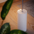 Witte cilindrische vloerlamp met slanke metalen staander en marmeren voet op een houten vloer tussen groene planten van sackit