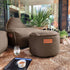 Bruine outdoor poef als voetensteun naast een lounge fauteuil in een lichte woonkamer met planten van sackit###
