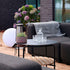 Ronde zwarte tuinsalontafel met teakhouten onderplank naast een loungesofa op een houten terras met glazen en een plantenpot in een sfeervolle buitenruimte