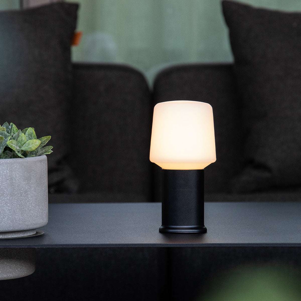 Zwarte sackit lampvoet met zacht oplichtende lampenkap op een tafel naast een plant en donkere kussens voor sfeervolle binnenverlichting