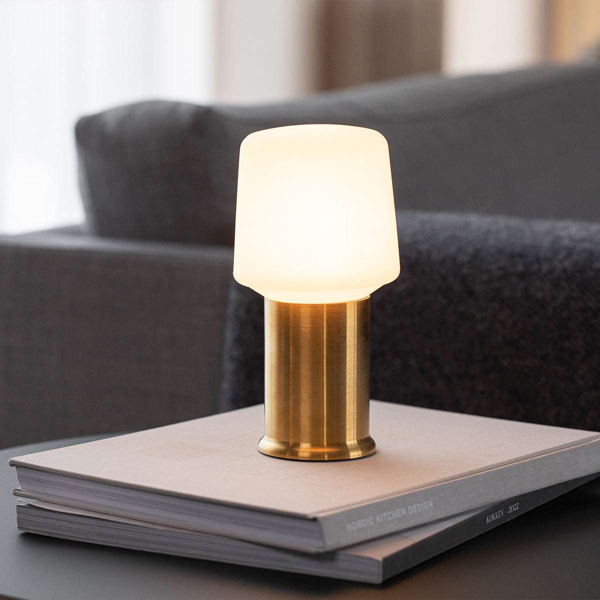 Ambience lampvoet in goudkleurig metaal met een zacht verlichte witte kap op een stapel boeken in een stijlvol interieur van sackit