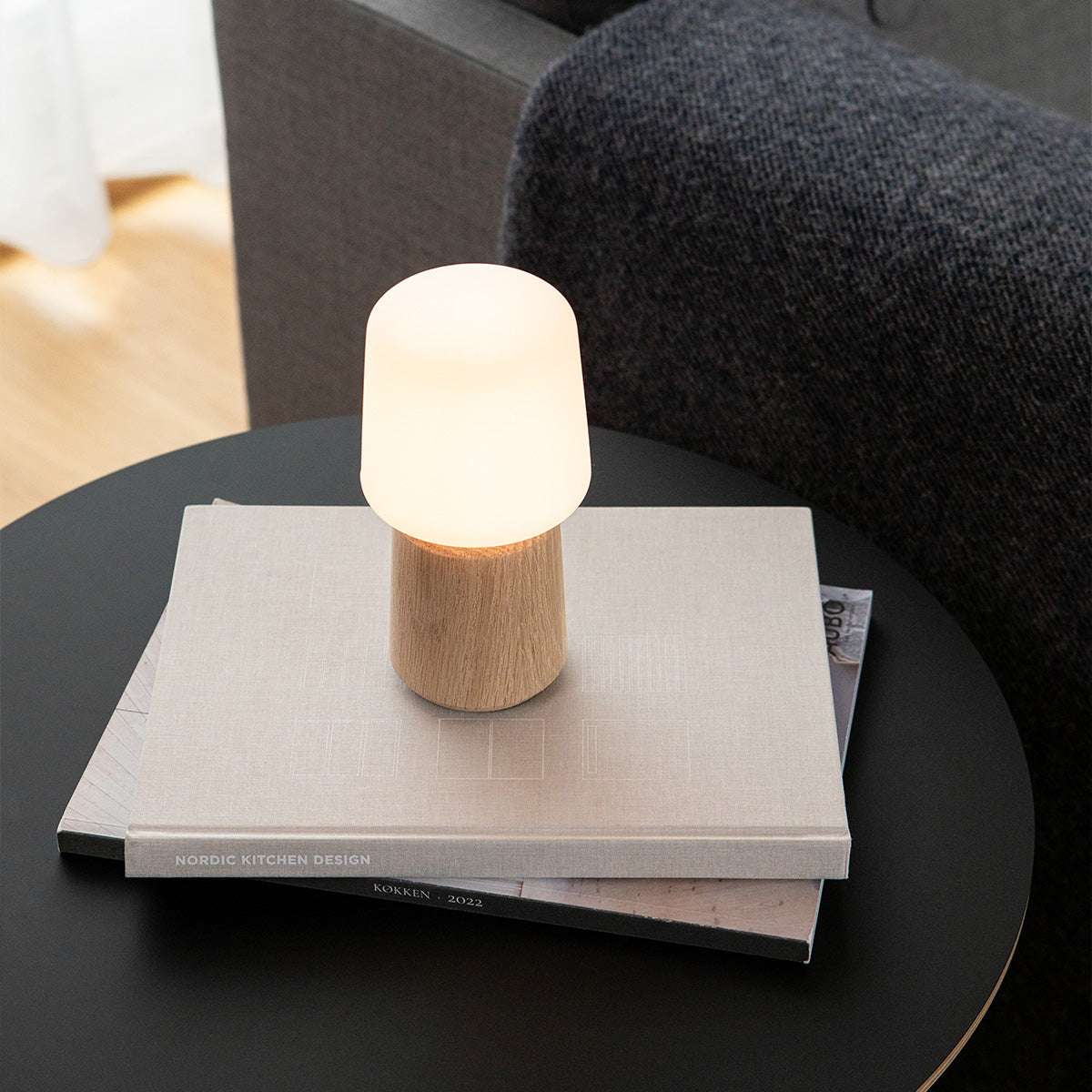Draadloze tafellamp met massief eikenhouten lampvoet en zacht wit licht op een stapel boeken op een ronde zwarte tafel van sackit