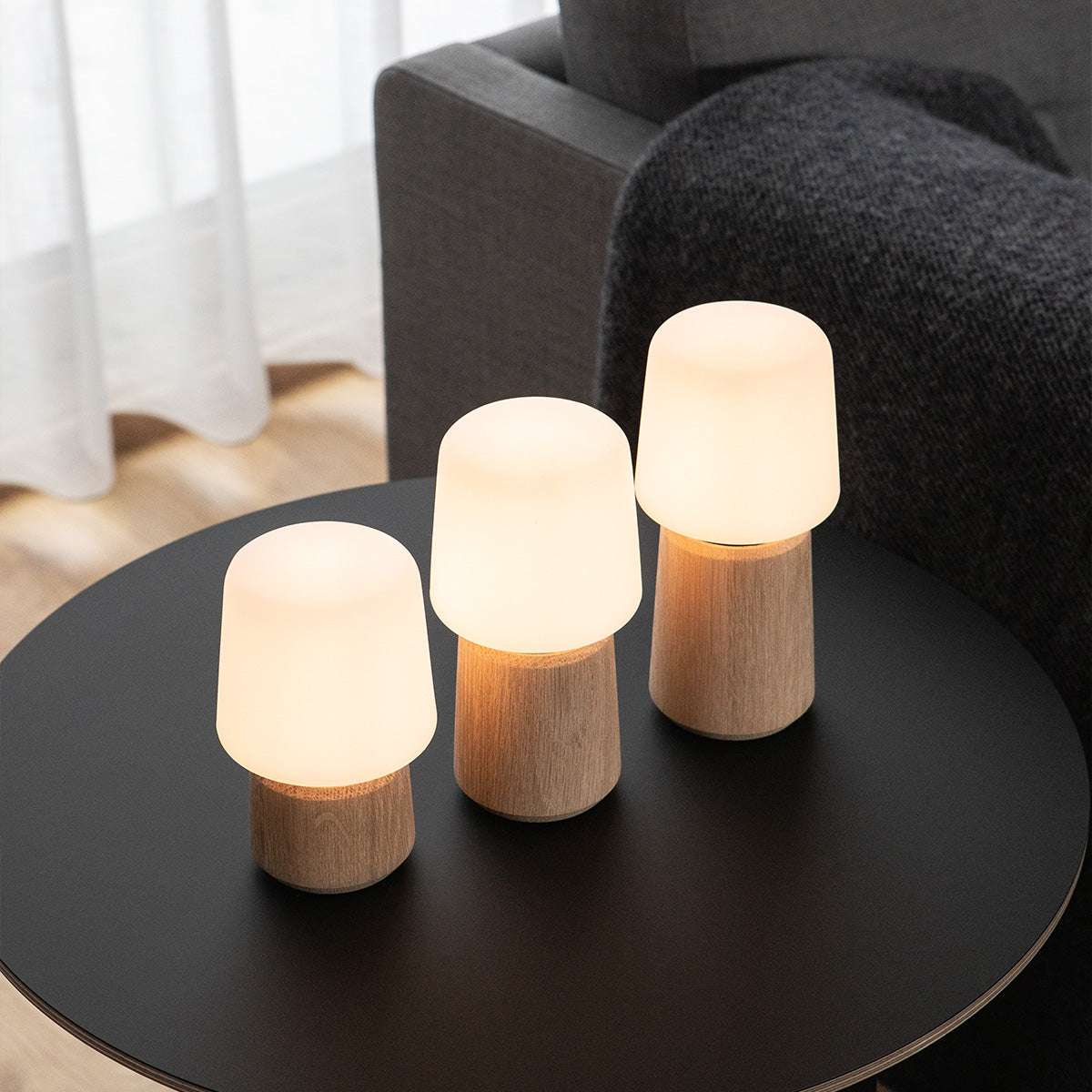 Drie draadloze tafellampen van sackit met warme verlichting en houten voet op een ronde zwarte tafel naast een comfortabele bank in een modern interieur