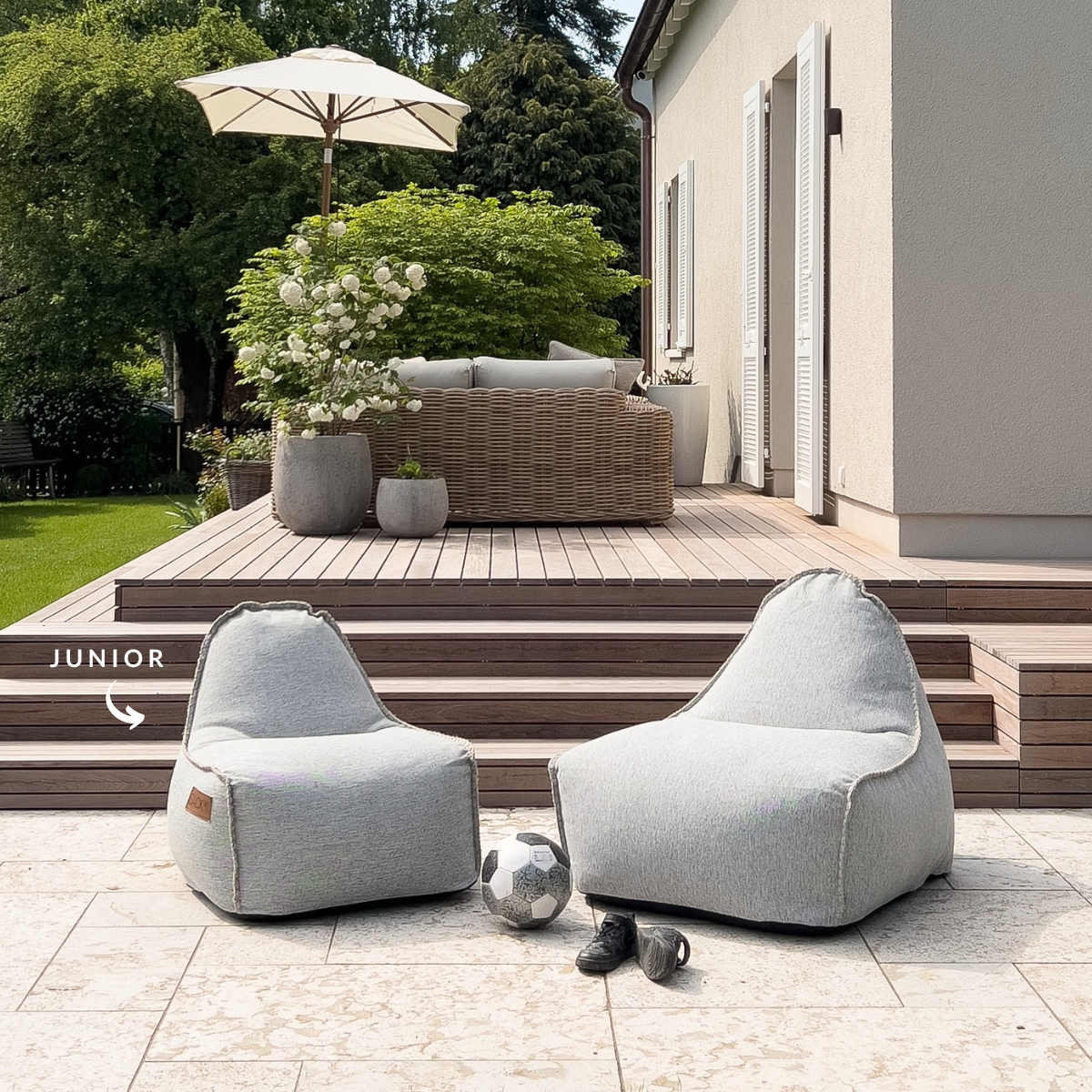 Twee lichtgrijze Cobana Junior zitzakstoelen van sackit op een zonnig terras met tuin en speelgoed voor comfortabel buiten zitten voor kinderen