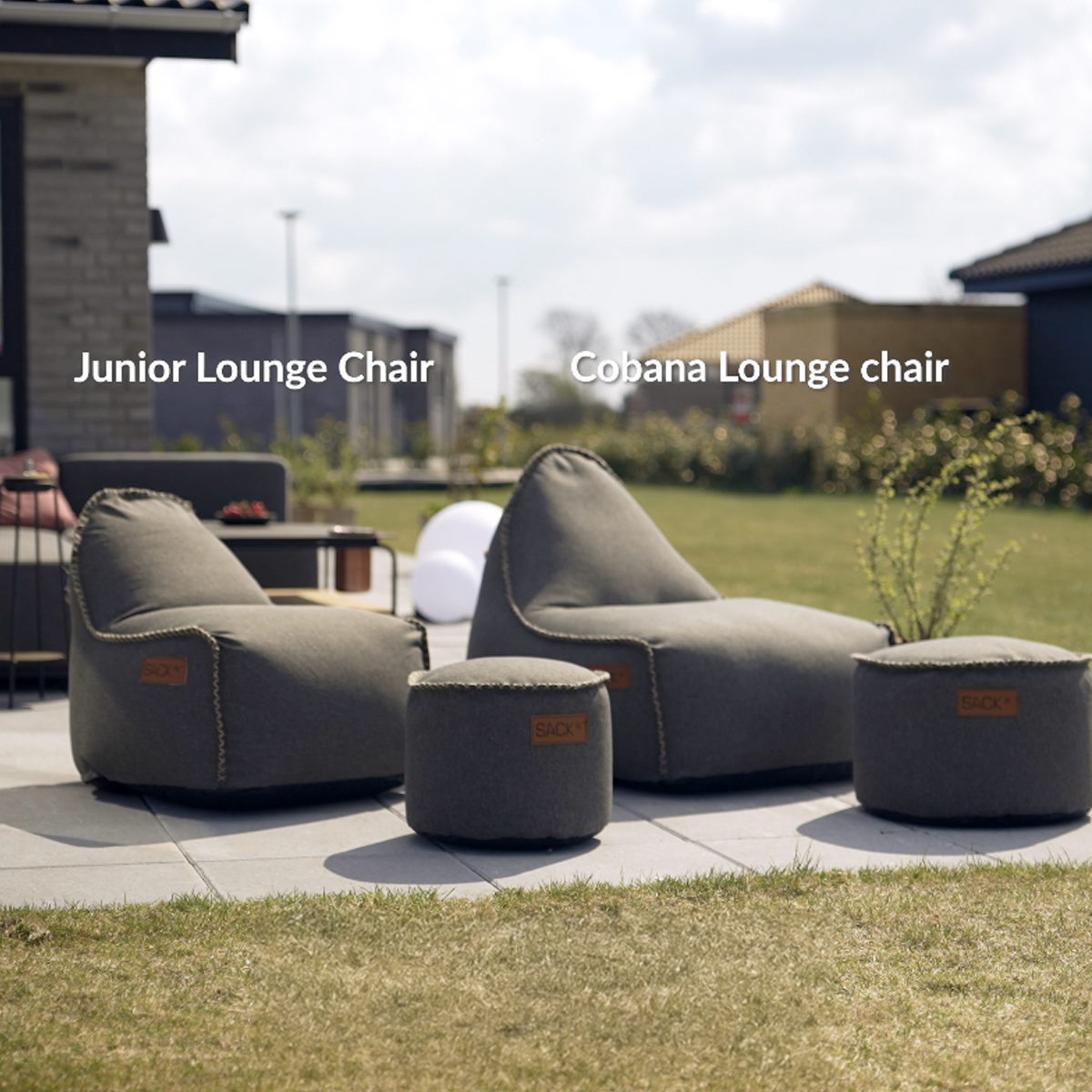 Donkergrijze junior lounge stoel en bijpassende poefs van sackit op een terras in een zonnige tuin met gras en moderne woningen op de achtergrond