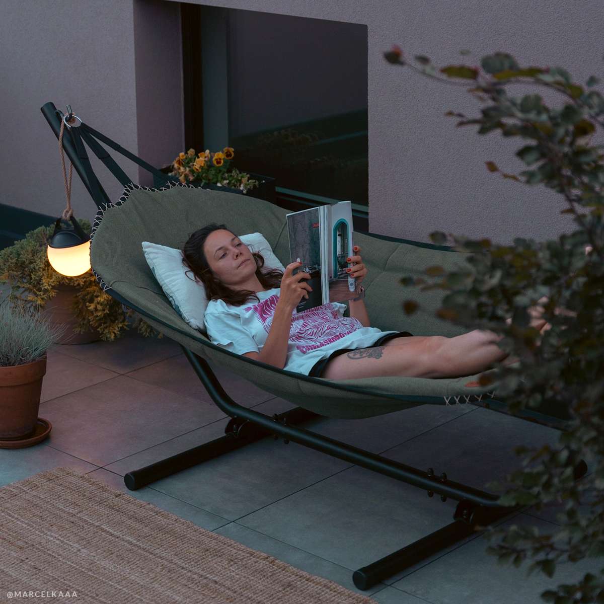 Vrouw leest ontspannen in een groene buitenhangmat van sackit op een terras bij avondlicht met kussens en een sfeervolle lamp naast zich