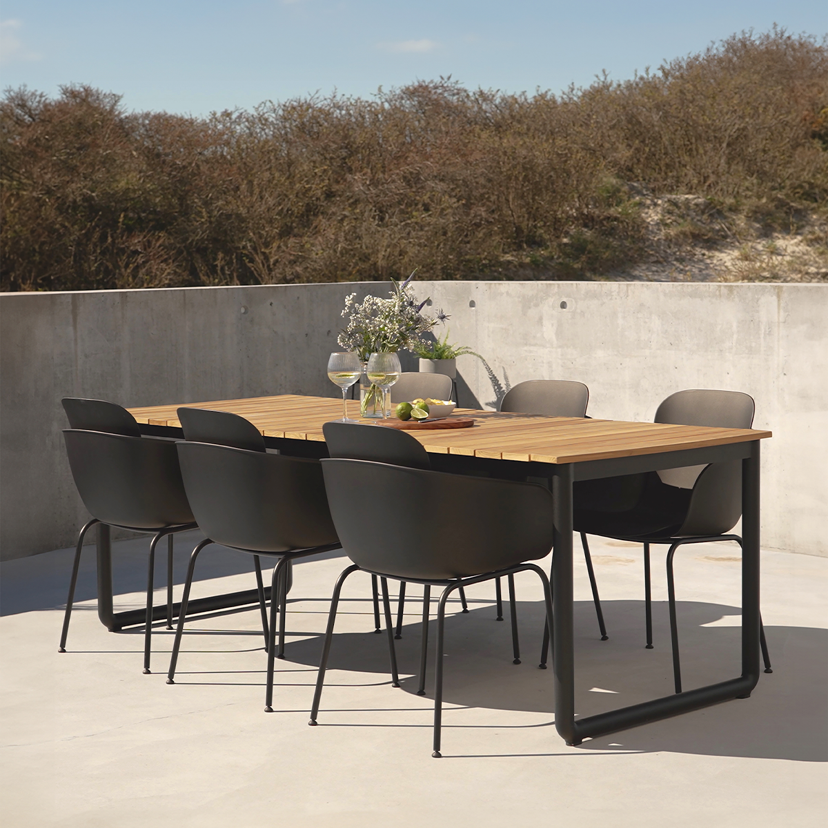 sackit buiten eettafel van hout met zes zwarte tuinstoelen op een zonnig terras bij een betonnen muur ontworpen voor nordisch buitenleven