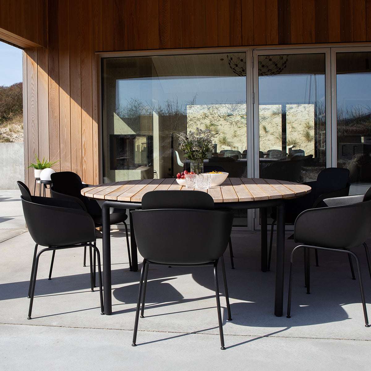 Zwarte patio eetstoelen van sackit rond een lichte houten tuintafel op een zonnig terras bij een moderne houten woning voor comfortabel buiten eten