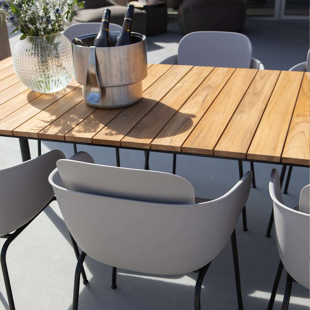 Grijze buitenstoelen rond een houten tuintafel met champagnekoeler en vaas op een zonnig terras van sackit ervaren design voor het buitenleven