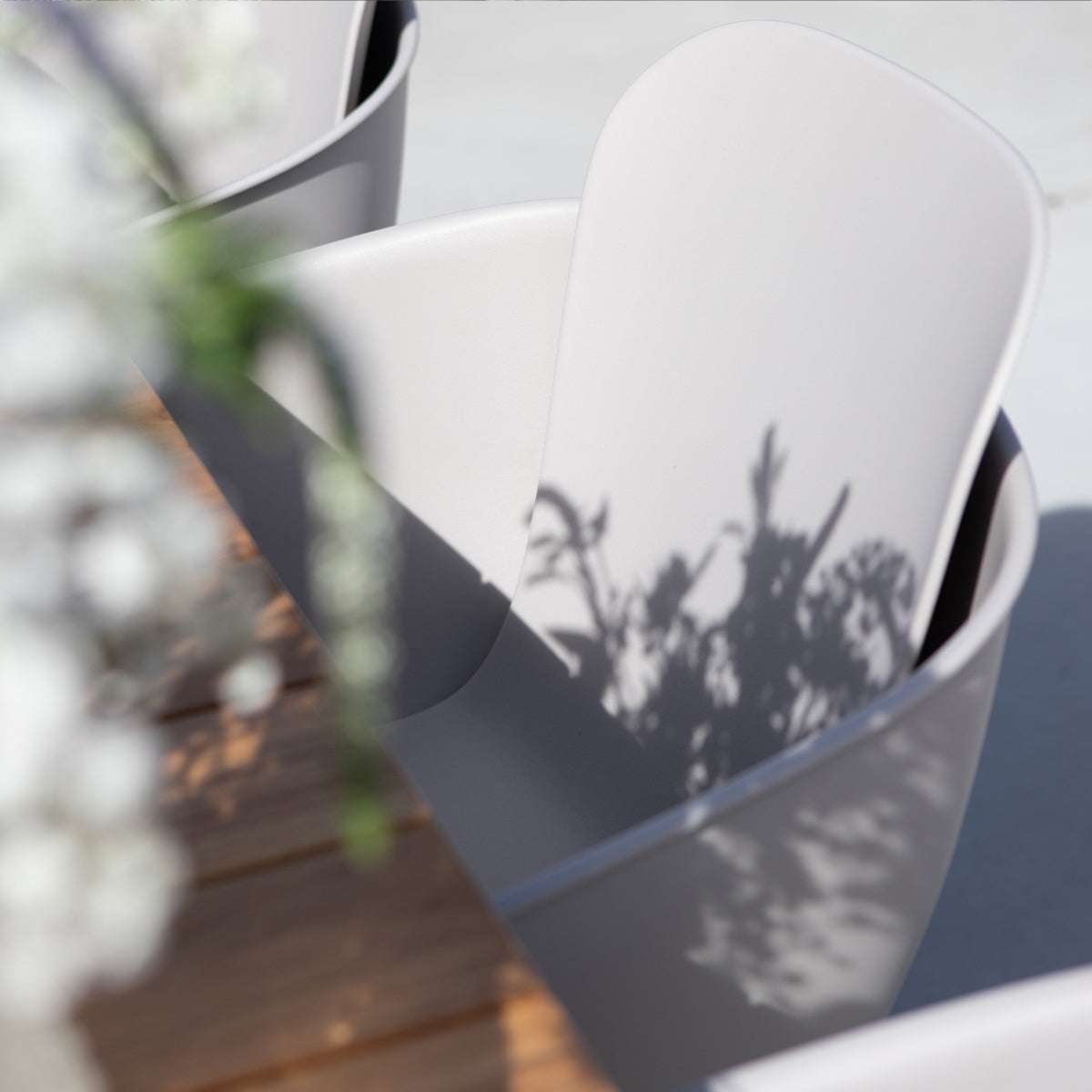 Witte outdoor eetkamerstoel met omarmende rugleuning op een houten terras in zonlicht met schaduwen van planten voor een rustige Scandinavische uitstraling
