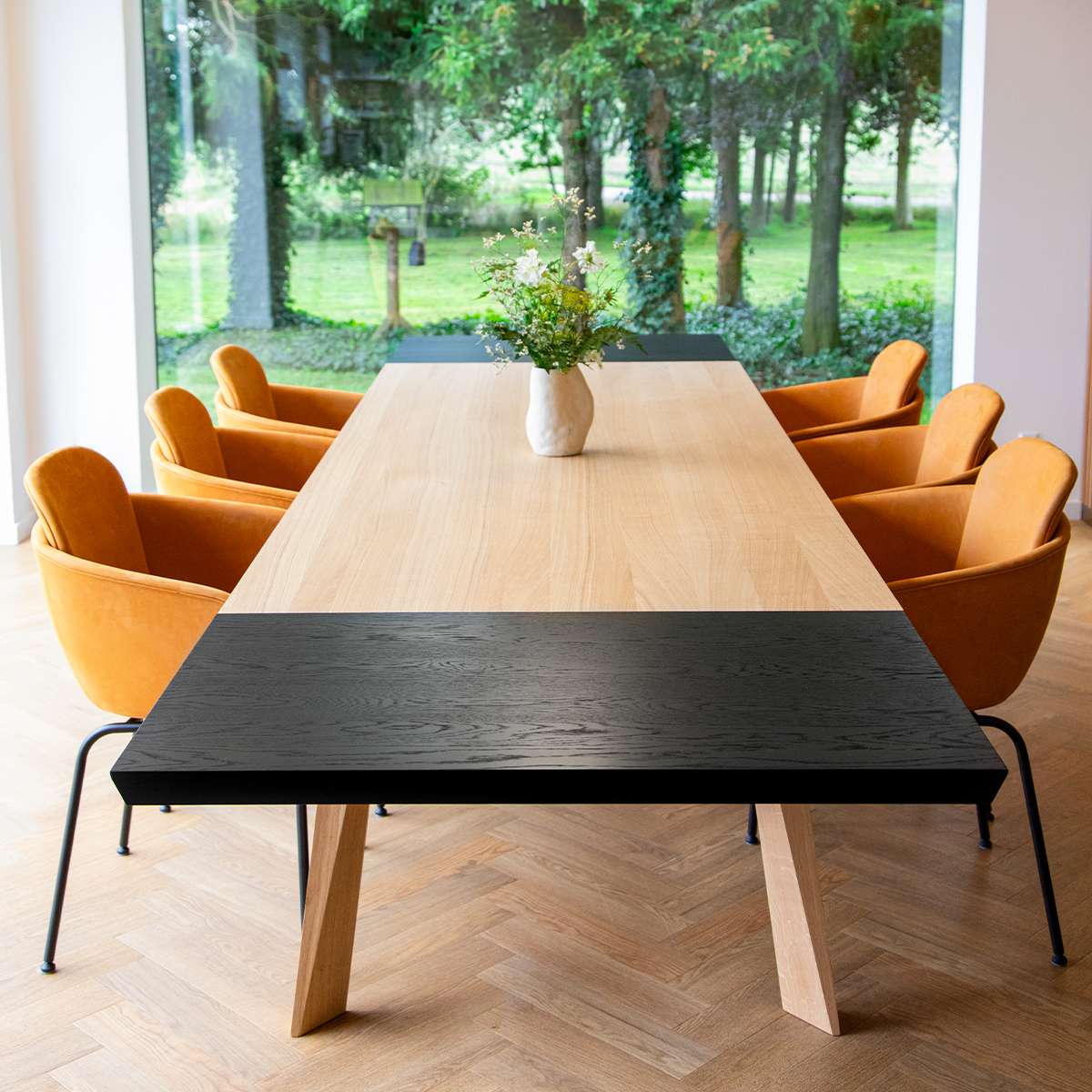 Lange houten eettafel met zwart gebeitst verlengblad en poten van licht hout omringd door elegante oranje eetkamerstoelen in een lichte ruimte met uitzicht op de tuin