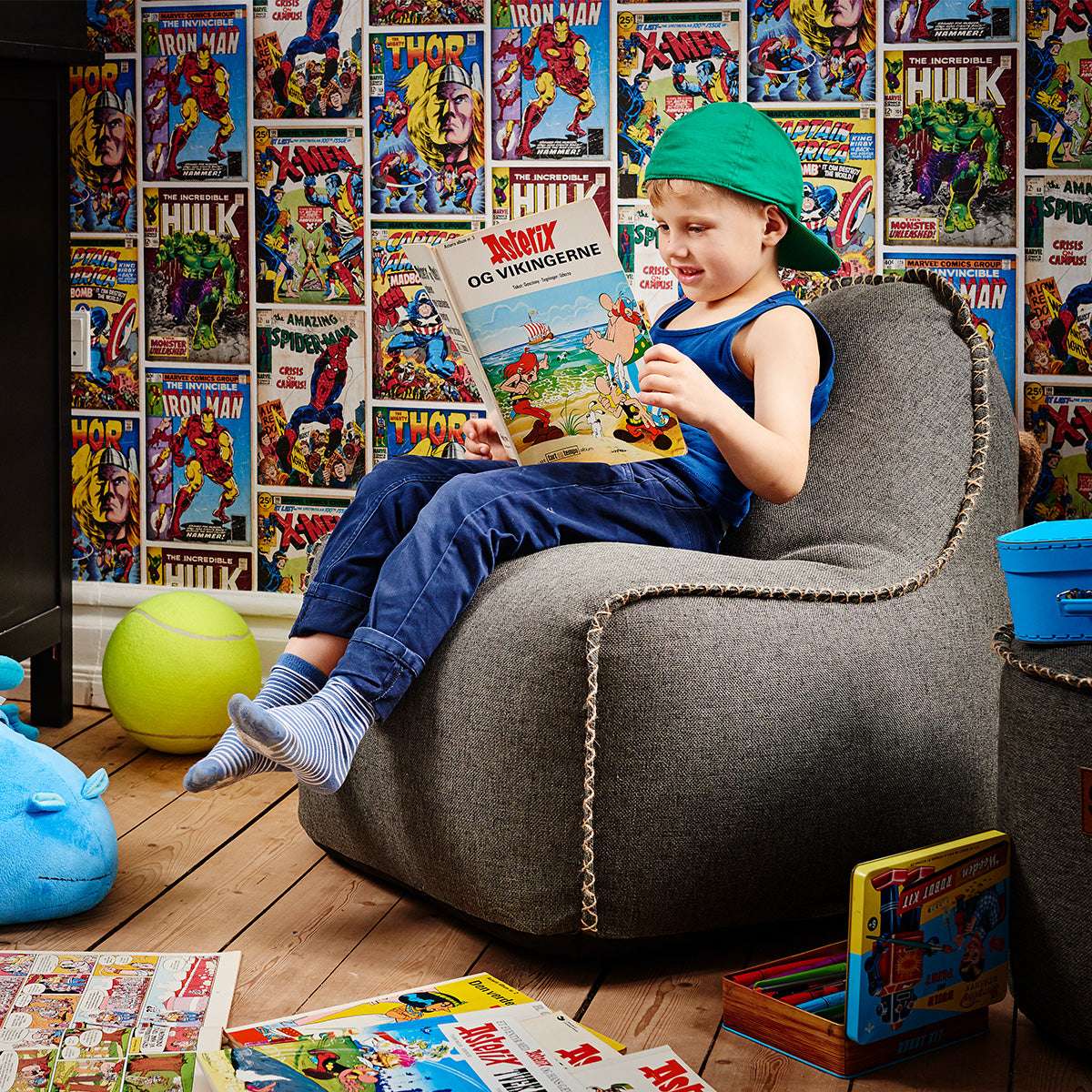 Kind leest een stripboek in een comfortabele grijze kinderzitzakstoel van sackit in een kleurrijke kamer met superheldenbehang en speelgoed
